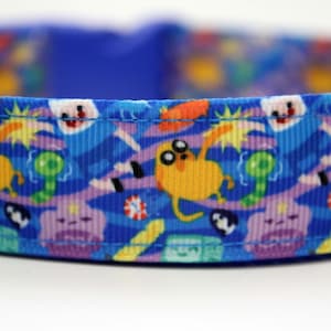 May include: Blue dog collar featuring a repeating pattern of cartoon characters in yellow, white, orange, and purple. The collar has a blue plastic buckle and is made of a durable, patterned fabric. The collar is designed for pets.