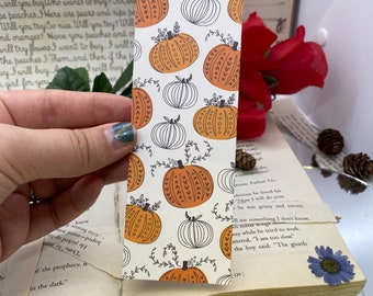 May include: A rectangular bookmark with a repeating pattern of orange and white pumpkins on a cream background. The pumpkins have decorative details and are surrounded by black vines. The bookmark is held over an open book.