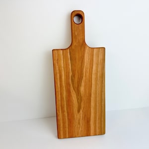 May include: A rectangular wooden cutting board with a handle and a hole for hanging. The board is made of light brown wood with visible grain patterns. The handle is rectangular with a rounded top.