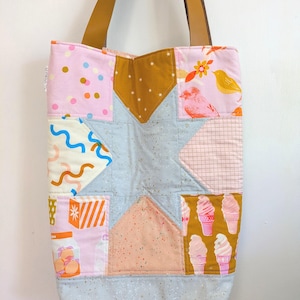 May include: A patchwork tote bag with a star design. The bag features various patterned fabrics in pink, blue, gold, and orange, with brown leather handles. The patterns include polka dots, birds, ice cream cones, and geometric shapes.