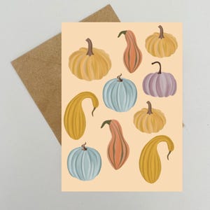 Puede incluir: Tarjeta de felicitación con ilustraciones de varias calabazas en amarillo, naranja, azul y morado. Las calabazas están dispuestas sobre un fondo melocotón claro. Se ve un sobre marrón en el fondo.