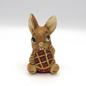 May include: A small brown rabbit figurine wearing a red dress with white polka dots and holding a pie in its paws.