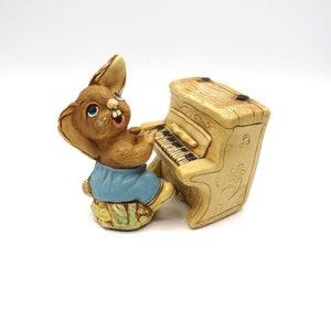 May include: A brown rabbit figurine wearing blue dungarees plays a small, beige piano with musical notes on the side. The figurine is made of ceramic and has a whimsical, cartoonish style.