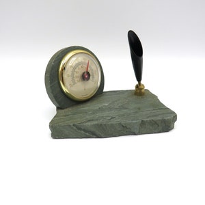 Könnte beinhalten: Ein Schiefer-Schreibtisch-Organizer mit einem Thermometer und einem Stifthalter. Das Thermometer hat einen goldenen Rand und ein weißes Zifferblatt mit schwarzen Markierungen. Der Stifthalter ist schwarz und hat eine Messingbasis.