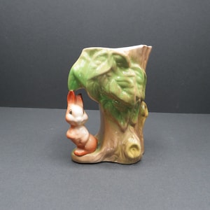 Könnte beinhalten: Eine Keramikvase in Form eines Baumstamms mit grünen Blättern und einem Eichhörnchen, das hinter dem Stamm hervorlugt.