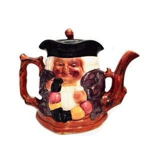 Op de afbeelding: Een theepot van keramiek in de vorm van een man met een zwarte hoed en een paarse mantel. De theepot heeft een bruine handgreep en tuit en is versierd met kleurrijke details.