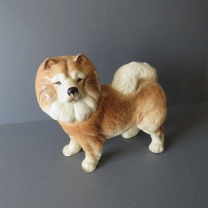 Op de afbeelding: Een keramiek beeldje van een bruine en witte Chow Chow hond. De hond staat met zijn hoofd schuin en zijn tong uitstekend.
