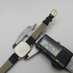 K&ouml;nnte beinhalten: Eine silberne Uhr mit einem schwarzen Lederarmband. Das Zifferblatt der Uhr ist quadratisch und das Armband ist 24,57 mm breit. Die Uhr tr&auml;gt den Text "HARVEY CALE MERCIAN" auf der R&uuml;ckseite.