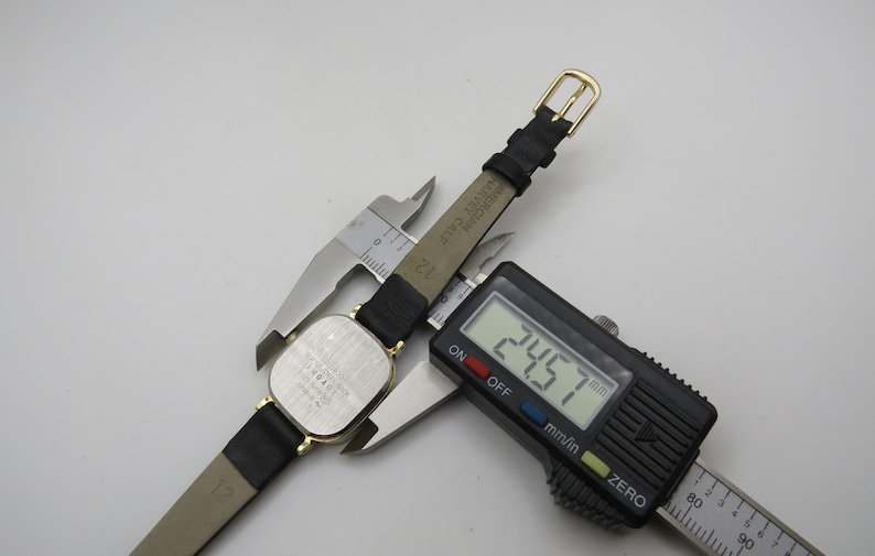 K&ouml;nnte beinhalten: Eine silberne Uhr mit einem schwarzen Lederarmband. Das Zifferblatt der Uhr ist quadratisch und das Armband ist 24,57 mm breit. Die Uhr tr&auml;gt den Text "HARVEY CALE MERCIAN" auf der R&uuml;ckseite.