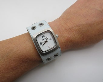 May include: A white Nixon wristwatch with a square face and a light blue leather band with silver eyelets. The watch face has black hour and minute hands and black Roman numeral hour markers.