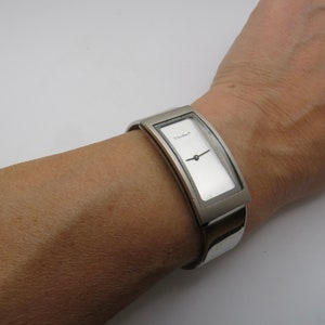 Puede incluir: Un reloj plateado con una esfera rectangular y una correa plateada. La esfera del reloj es blanca con agujas negras y la palabra "Ebel" impresa en ella.