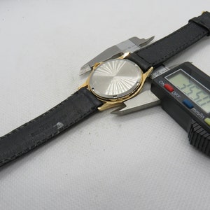 May include: A gold-tone wristwatch with a black leather band. The watch face is silver with a sunburst pattern. The watch is being measured with a digital caliper, which reads 35.27 mm.