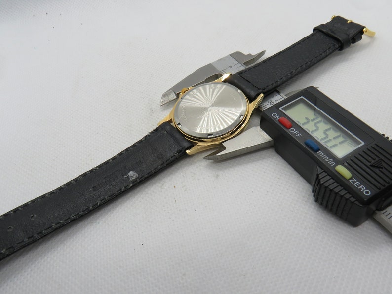 May include: A gold-tone wristwatch with a black leather band. The watch face is silver with a sunburst pattern. The watch is being measured with a digital caliper, which reads 35.27 mm.