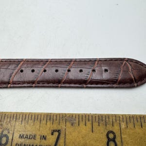 May include: Close-up of a brown leather watch strap with a crocodile skin texture. The strap has several holes for adjustment. A yellow ruler with the text "MADE IN DENMARK" is below the strap.