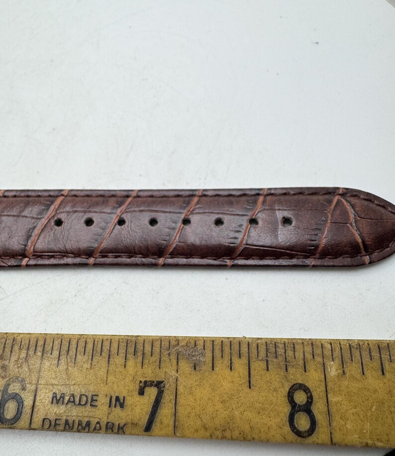 May include: Close-up of a brown leather watch strap with a crocodile skin texture. The strap has several holes for adjustment. A yellow ruler with the text "MADE IN DENMARK" is below the strap.