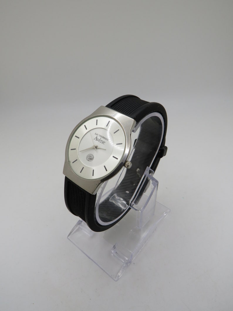 May include: A silver wristwatch with a white face and black rubber band. The watch face has the text "Eau Thermale Av&egrave;ne" printed on it.