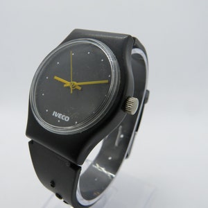 K&ouml;nnte beinhalten: Schwarze Armbanduhr mit rundem Zifferblatt und gelben Zeigern. Auf dem Zifferblatt der Uhr steht das Wort "IVECO". Das Armband ist schwarz und scheint aus Gummi oder Kunststoff zu sein.
