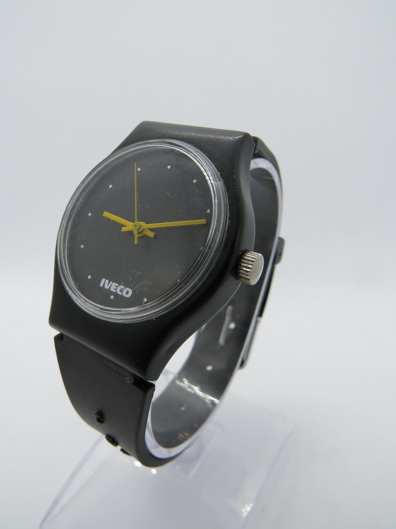 K&ouml;nnte beinhalten: Schwarze Armbanduhr mit rundem Zifferblatt und gelben Zeigern. Auf dem Zifferblatt der Uhr steht das Wort "IVECO". Das Armband ist schwarz und scheint aus Gummi oder Kunststoff zu sein.