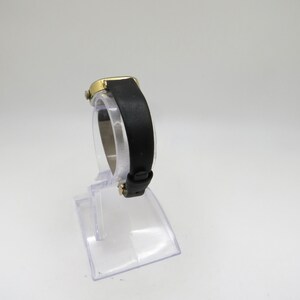 K&ouml;nnte beinhalten: Eine goldfarbene Armbanduhr mit einem schwarzen Lederarmband. Die Uhr befindet sich auf einem durchsichtigen Kunststoffst&auml;nder.