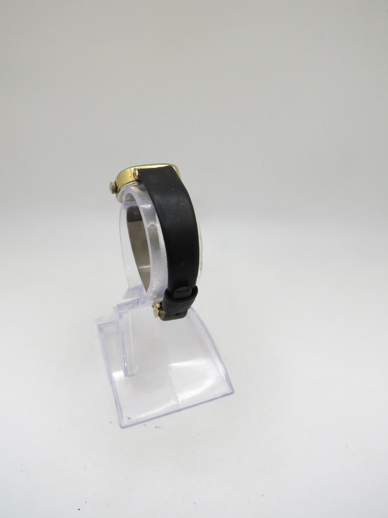K&ouml;nnte beinhalten: Eine goldfarbene Armbanduhr mit einem schwarzen Lederarmband. Die Uhr befindet sich auf einem durchsichtigen Kunststoffst&auml;nder.