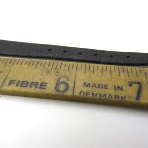 K&ouml;nnte beinhalten: Ein schwarzes Lederarmband, das mit einem Lineal gemessen wird. Das Lineal ist mit den Zahlen 5, 6 und 7 gekennzeichnet. Das Lineal tr&auml;gt au&szlig;erdem die Aufschrift "Fibre" und "Made in Denmark".