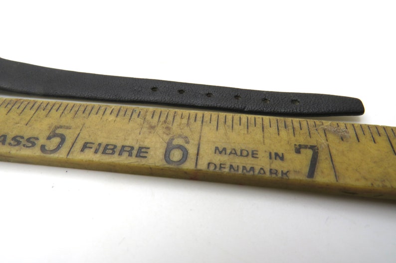 K&ouml;nnte beinhalten: Ein schwarzes Lederarmband, das mit einem Lineal gemessen wird. Das Lineal ist mit den Zahlen 5, 6 und 7 gekennzeichnet. Das Lineal tr&auml;gt au&szlig;erdem die Aufschrift "Fibre" und "Made in Denmark".