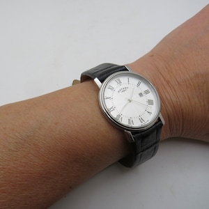 May include: A silver Rotary wristwatch with a white face, black leather band, and Roman numeral hour markers. The watch has a date window at the 3 o'clock position.