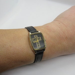 May include: A black leather wristwatch with a square gold-tone face featuring a gold cross with black floral details.