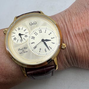 May include: A gold-toned wristwatch with a cream-colored face and a brown leather band. The watch features two dials, one with Roman numerals and the other with Arabic numerals. The watch face has the text "Dual Time" and "WR 5BAR".