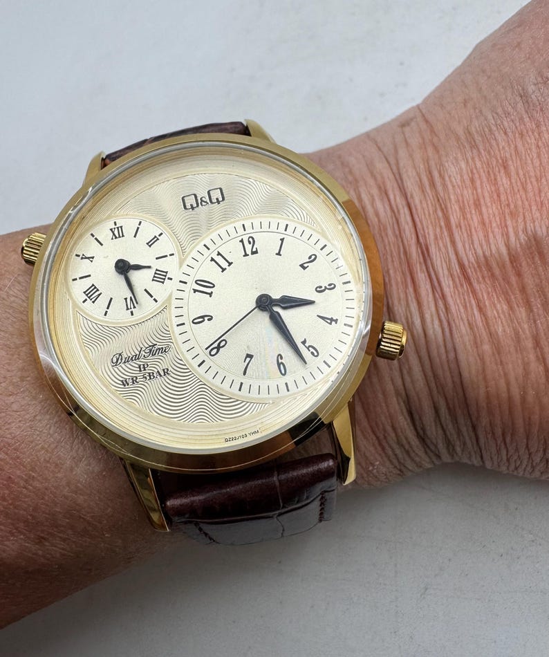 May include: A gold-toned wristwatch with a cream-colored face and a brown leather band. The watch features two dials, one with Roman numerals and the other with Arabic numerals. The watch face has the text "Dual Time" and "WR 5BAR".