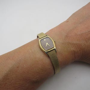 Puede incluir: Un reloj de pulsera dorado con una esfera rectangular y una correa de malla. La esfera del reloj es de color rosa apagado con agujas doradas y números romanos. El reloj está sujeto a una muñeca.