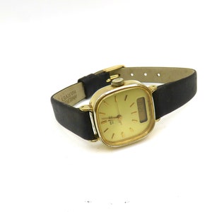 K&ouml;nnte beinhalten: Eine goldfarbene analoge Armbanduhr mit quadratischem Zifferblatt und schwarzem Lederarmband. Das Zifferblatt hat einen wei&szlig;en Hintergrund mit schwarzen Zeigern und einem Datumsfenster. Das Armband ist mit "AMER. HARVEY" beschriftet.