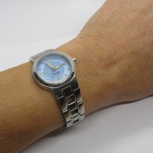 May include: A silver-toned wristwatch with a blue face and a silver band. The watch face has a white outline around the numbers and the word "roxy" in the center.