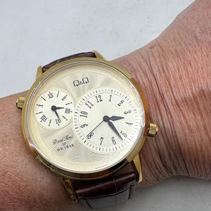 May include: Gold-toned dual-time wristwatch with a brown leather band. The watch face has two dials, one with Roman numerals and the other with Arabic numerals. The watch has the text "Dual Time WR 5BAR".