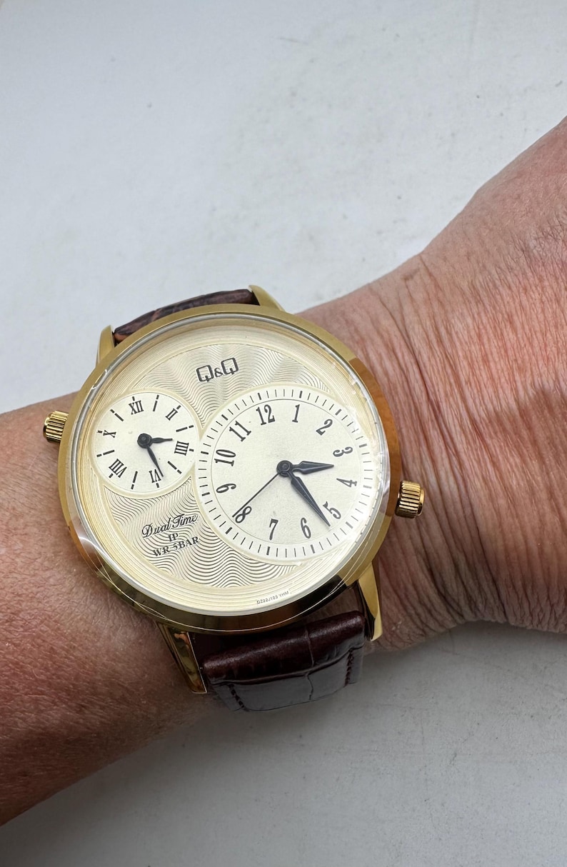 May include: Gold-toned dual-time wristwatch with a brown leather band. The watch face has two dials, one with Roman numerals and the other with Arabic numerals. The watch has the text "Dual Time WR 5BAR".