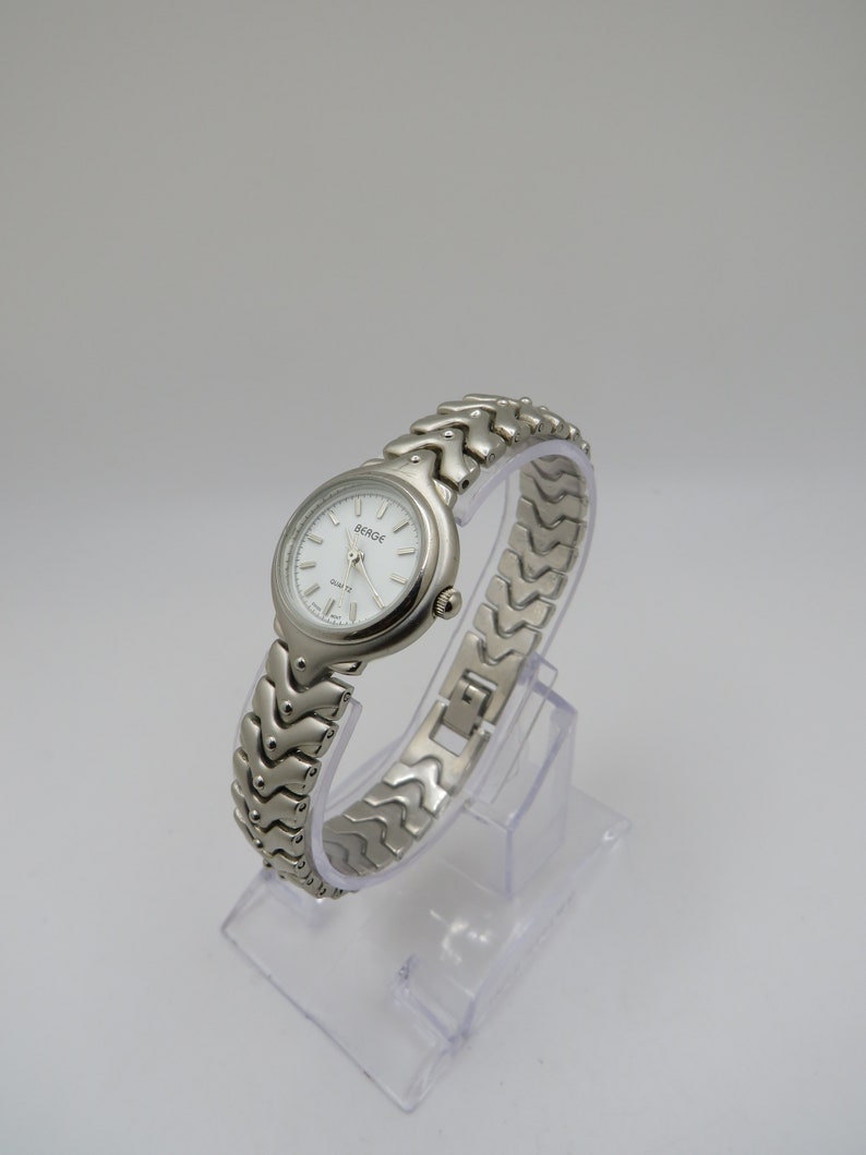 K&ouml;nnte beinhalten: Eine silberfarbene Armbanduhr mit wei&szlig;em Zifferblatt und silbernem Armband. Auf dem Zifferblatt der Uhr steht "BERGE" und "QUARTZ".