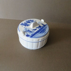 May include: A white ceramic jar with a blue and white Delftware design of windmills and houses. The jar has a lid with a knob on top.