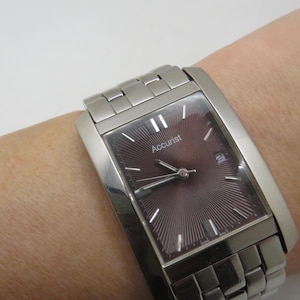 May include: A silver Accurist wristwatch with a rectangular face and a burgundy dial. The watch has a date display and a silver strap. The watch is on a wrist.