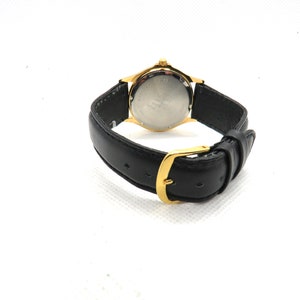 May include: A gold-tone watch with a black leather band. The watch face is silver and has black markings. The watch has a gold-tone buckle.