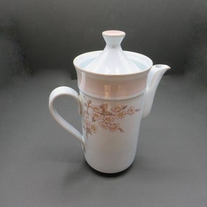 May include: White ceramic coffee pot with a pink and gold floral design. The pot has a lid and a handle.