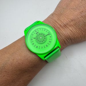 May include: A bright neon green wristwatch featuring a round case and matching strap. The dial displays standard hour numerals from 1 to 12, with smaller minute markers and half-hour increments printed in a circular pattern. The Softech logo is visible.