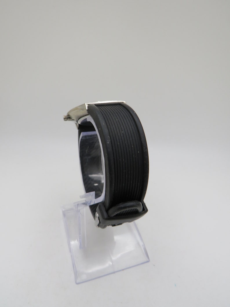 May include: A silver and black wristwatch with a black rubber band. The watch is on a clear display stand.