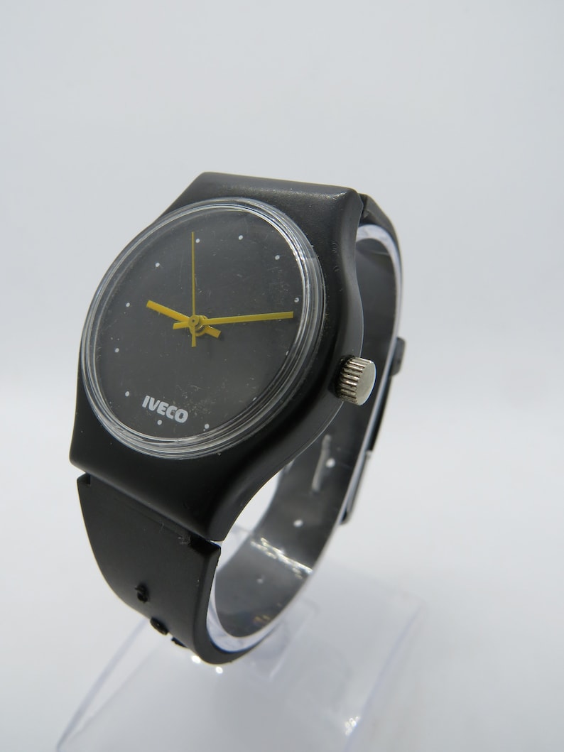 K&ouml;nnte beinhalten: Schwarze Armbanduhr mit rundem Zifferblatt und gelben Zeigern. Auf dem Zifferblatt der Uhr steht das Wort "IVECO". Das Armband ist schwarz und scheint aus Gummi oder Kunststoff zu sein.