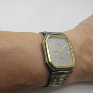 Könnte beinhalten: Eine silberne und goldfarbene Rotary-Uhr mit quadratischem Zifferblatt und einem zweifarbigen Metallband. Das Zifferblatt ist weiß mit schwarzen Zeigern und schwarzen römischen Ziffern als Stundenmarkierungen. Auf dem Zifferblatt steht außerdem "Rotary" und "30M Quartz".