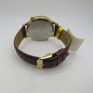 May include: Gold-tone wristwatch with a brown leather band. The watch has a round face and a buckle closure. The band has a crocodile pattern. A yellow tag is attached to the band.