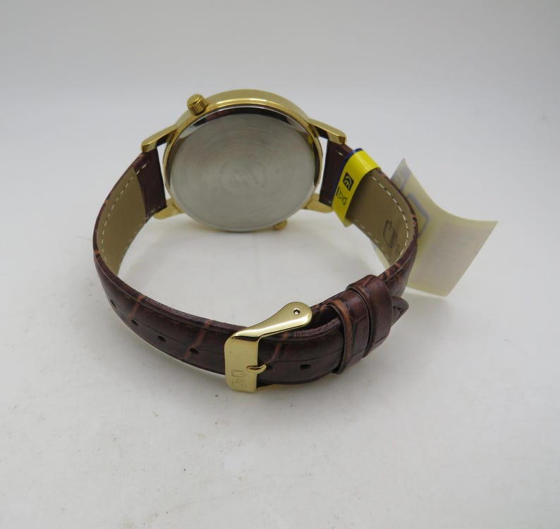 May include: Gold-tone wristwatch with a brown leather band. The watch has a round face and a buckle closure. The band has a crocodile pattern. A yellow tag is attached to the band.
