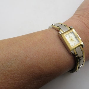 Könnte beinhalten: Eine zweifarbige Gold- und Silberuhr mit quadratischem Zifferblatt und weißem Zifferblatt. Die Uhr hat ein zweifarbiges Armband und einen kleinen Diamanten an der 12-Uhr-Position.