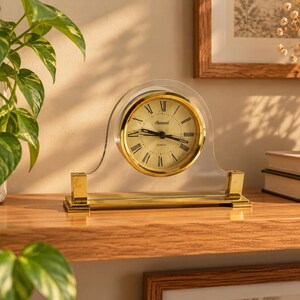 Puede incluir: Un reloj de sobremesa con esfera dorada circular, números romanos y manecillas negras, encerrado en un marco de acrílico transparente en forma de arco. El reloj se apoya sobre una base rectangular de metal en tono dorado.