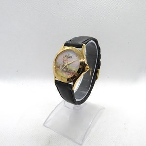 May include: A gold-tone wristwatch with a black leather band. The watch face features a white background with a painted scene of a village and a windmill. The watch face also has Roman numeral hour markers and the word "Village" printed on it.