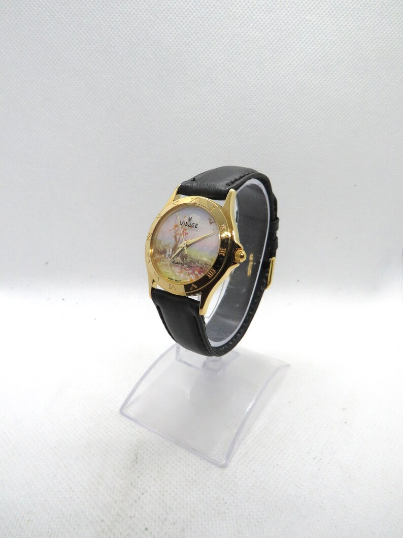 May include: A gold-tone wristwatch with a black leather band. The watch face features a white background with a painted scene of a village and a windmill. The watch face also has Roman numeral hour markers and the word "Village" printed on it.